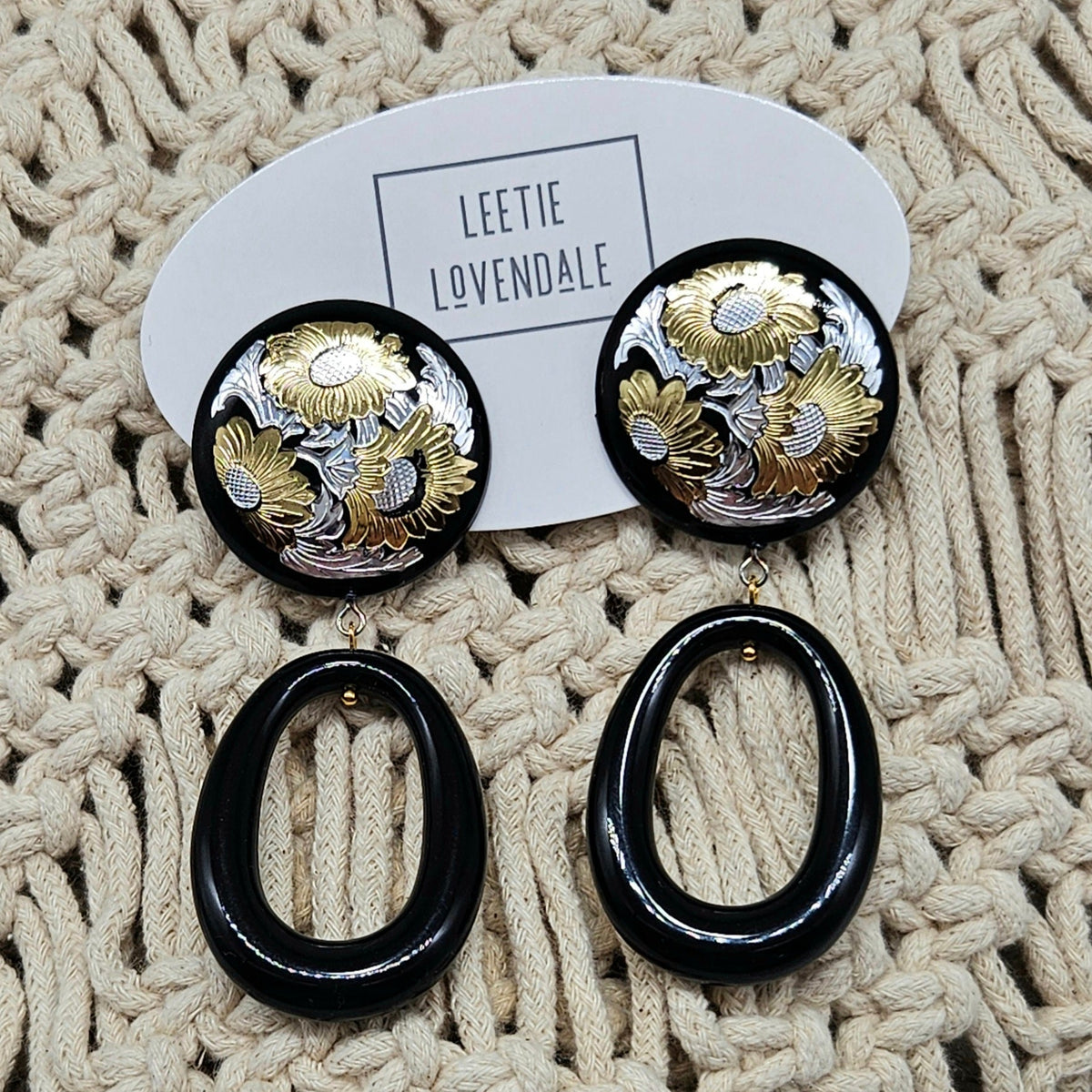 Pair of earrings with black and gold design on a textured beige surface, branded 'LEETIE LOVENDALE'.