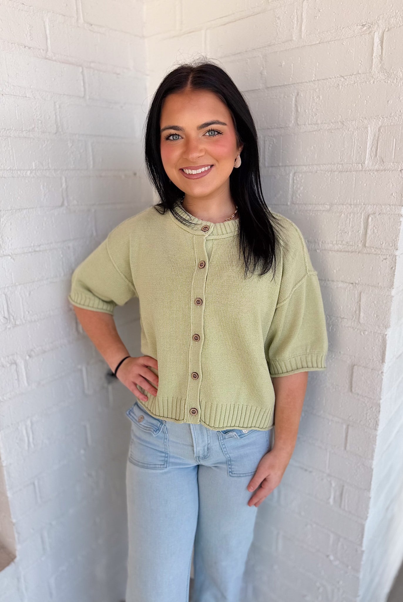 Woman wearing a light green knitted cardigan and light blue jeans against a white brick wall.