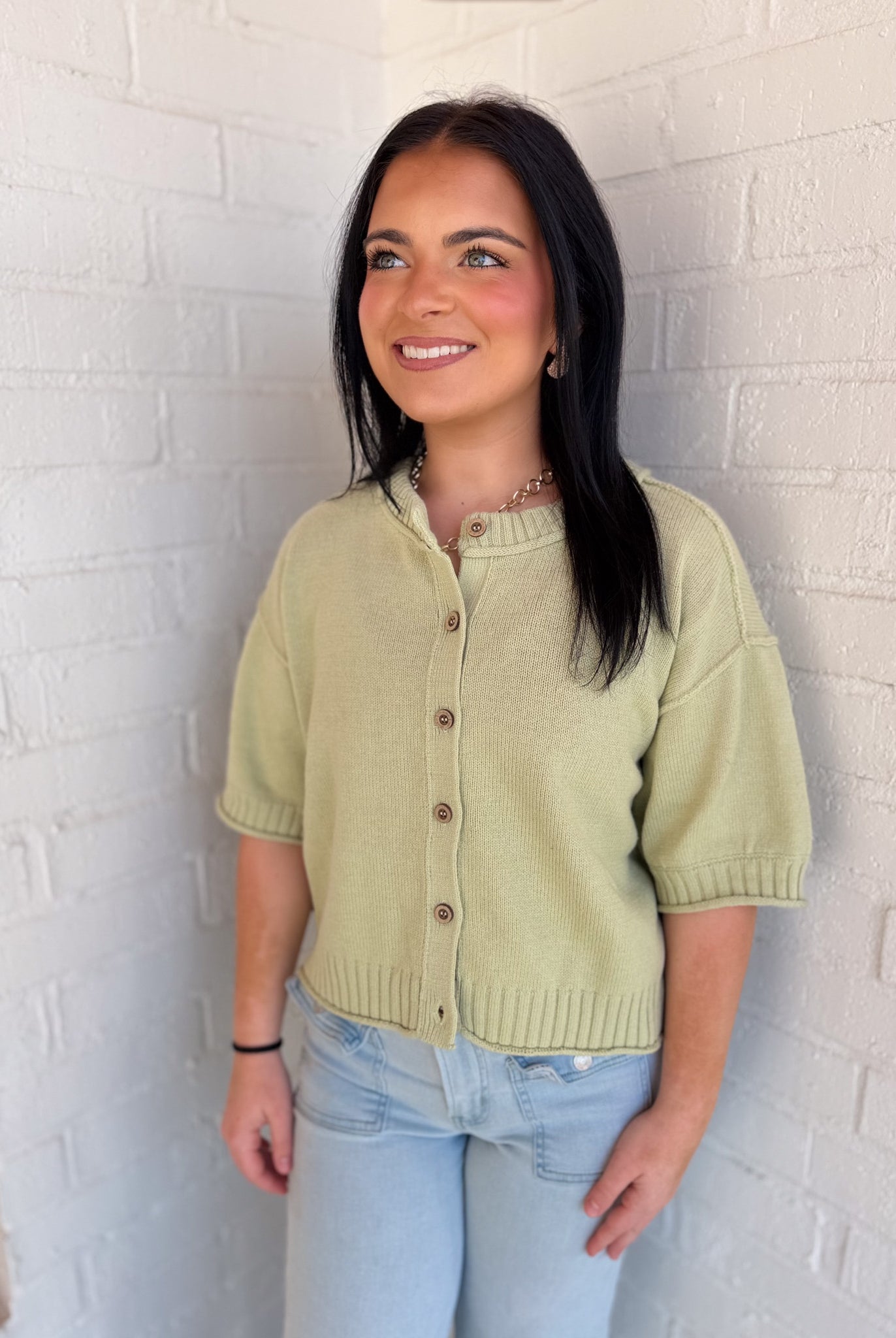 Woman wearing a light green short-sleeve cardigan against a white brick wall.