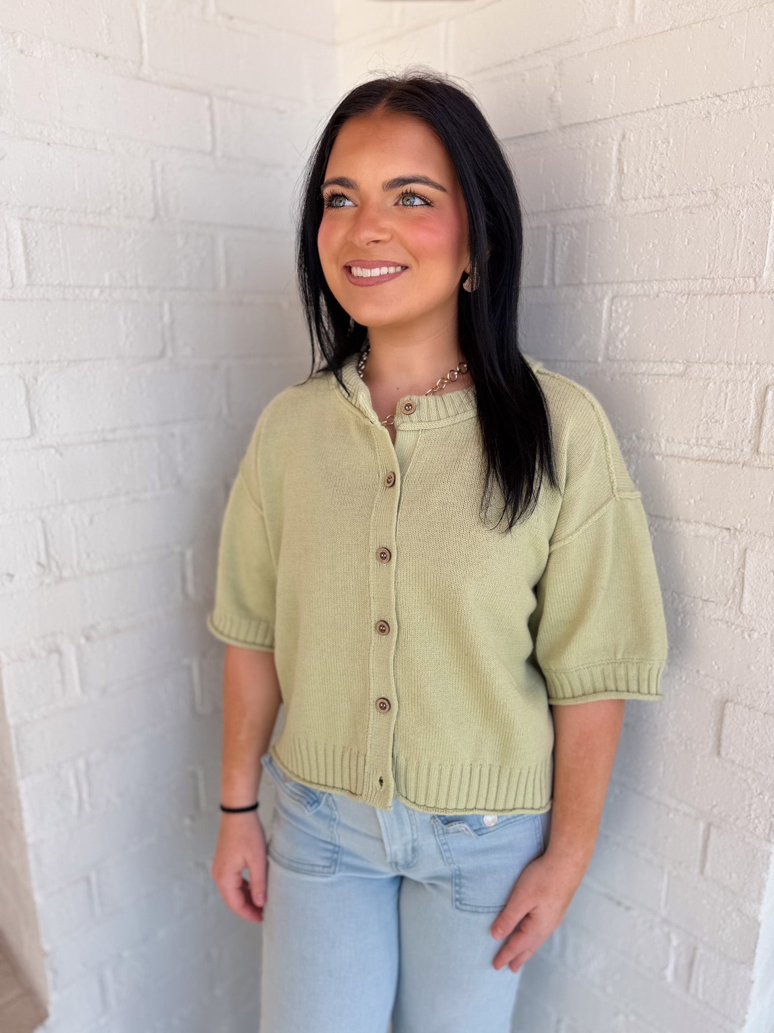 Woman wearing a light green short-sleeve cardigan against a white brick wall.