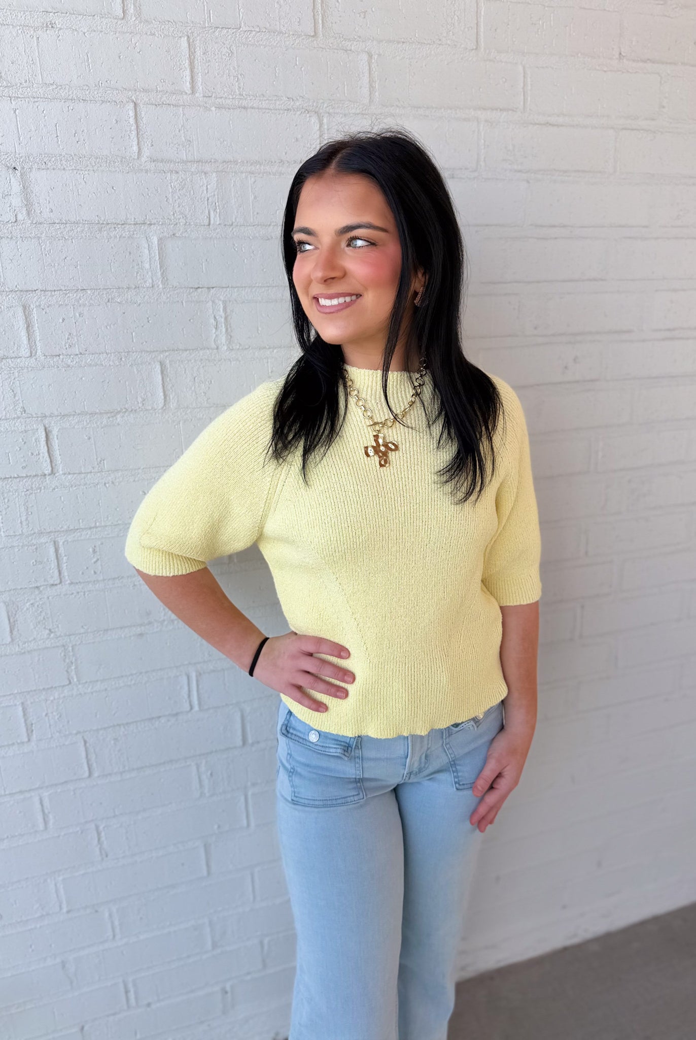 Woman wearing a yellow sweater and light blue jeans against a white brick wall.