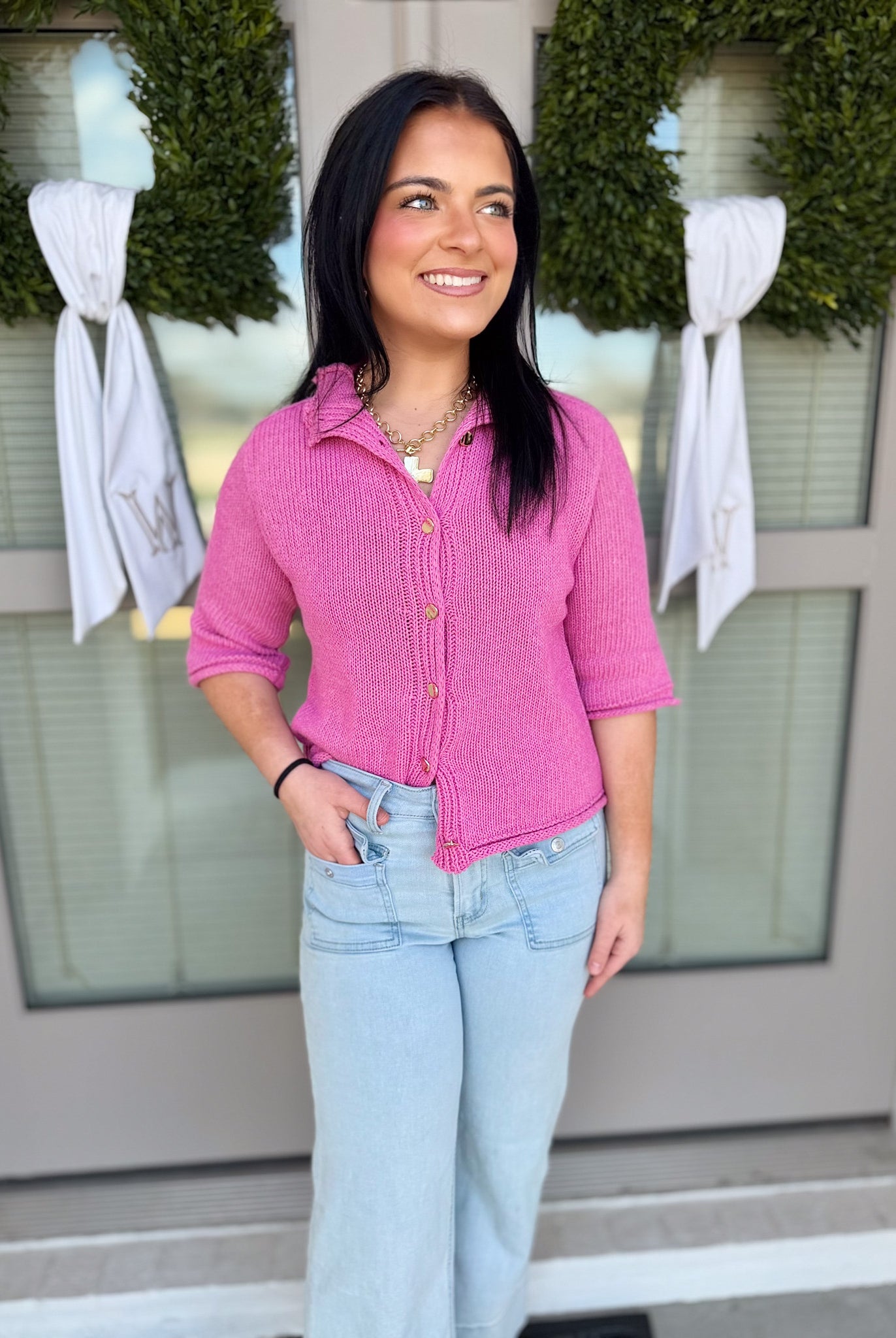 Woman wearing a pink shirt and light blue jeans standing in front of a door with wreaths and bows.
