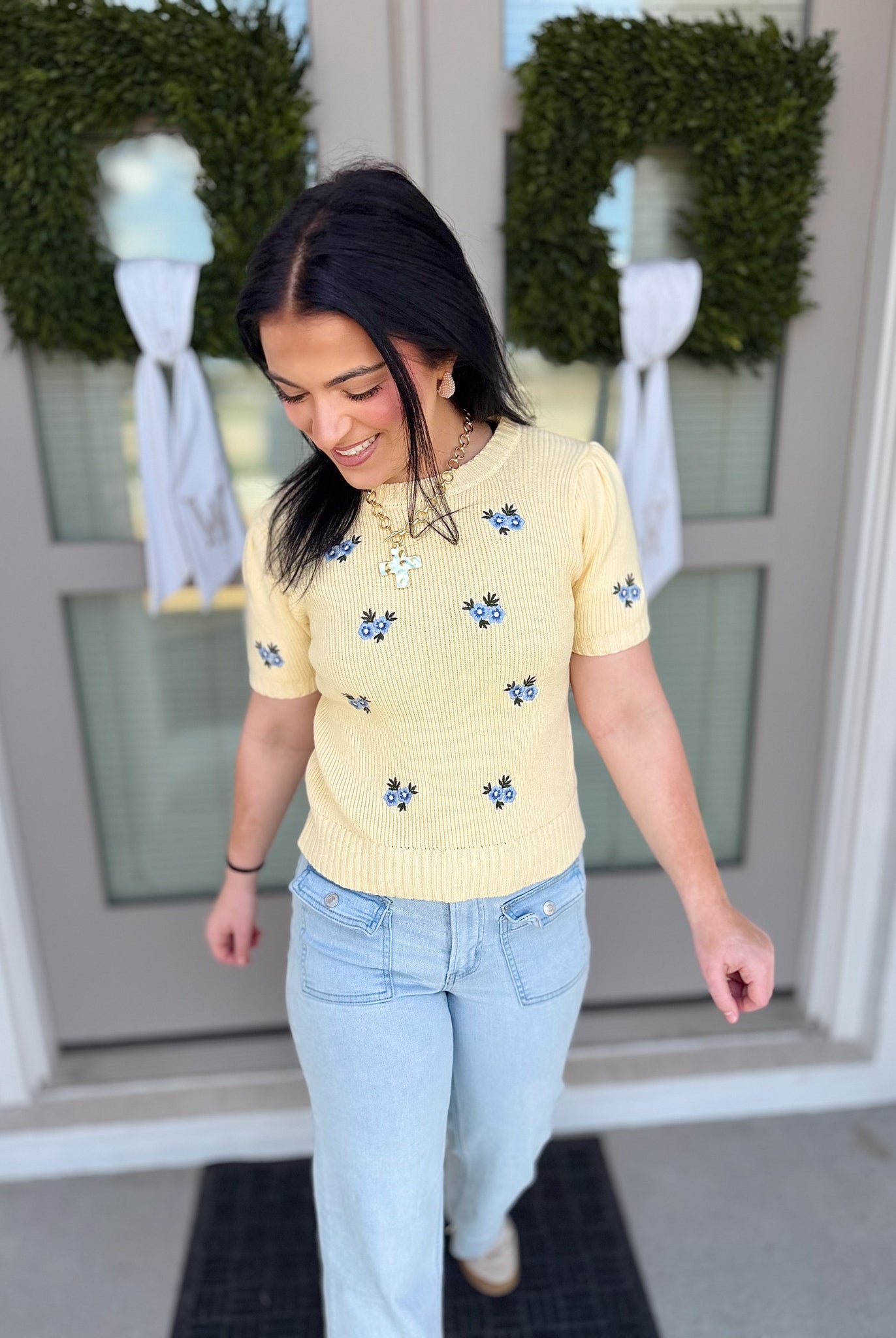Woman wearing a yellow floral sweater and light blue jeans standing in front of a door with wreaths.
