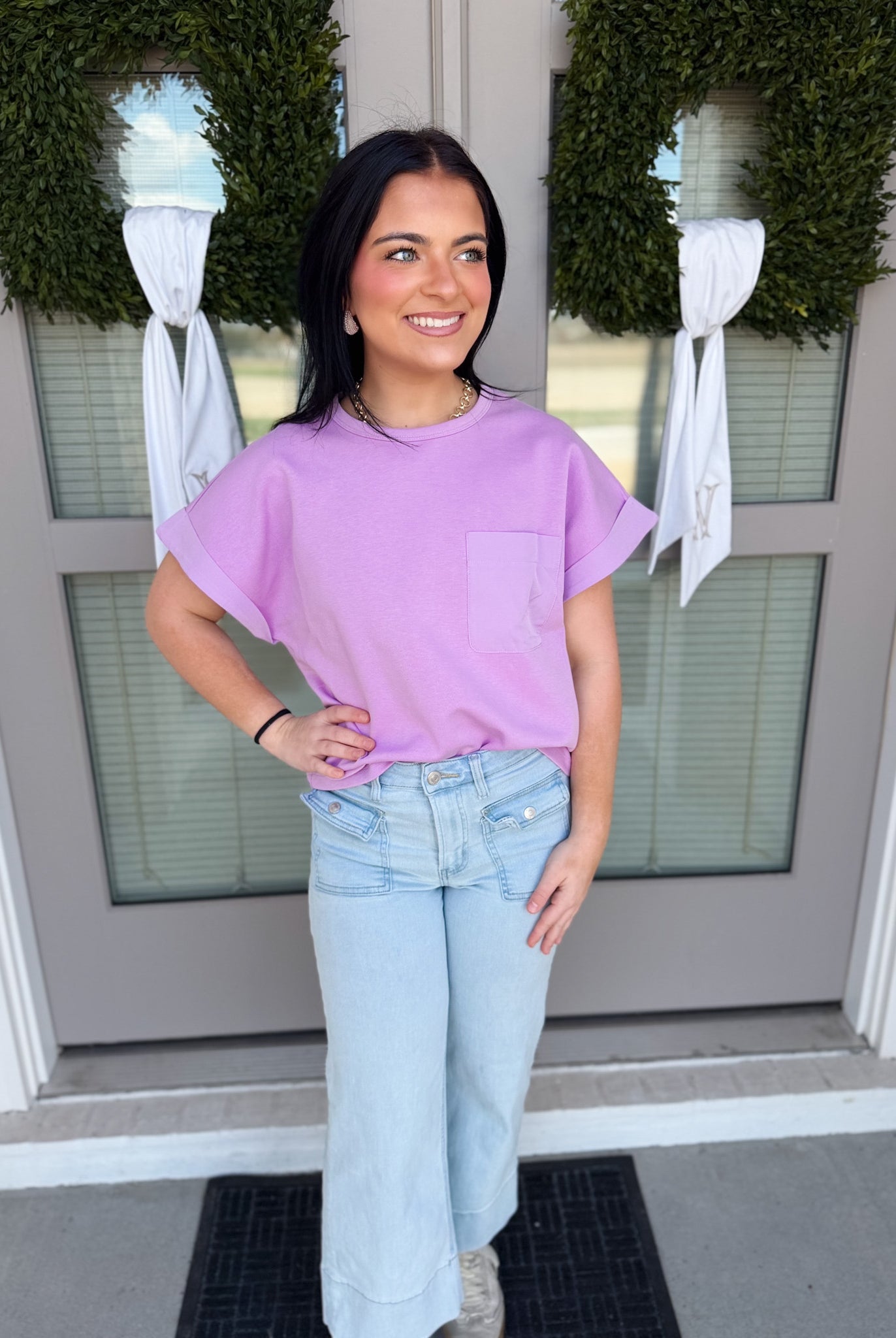 Woman wearing a purple shirt and light blue jeans standing in front of a door with wreaths.