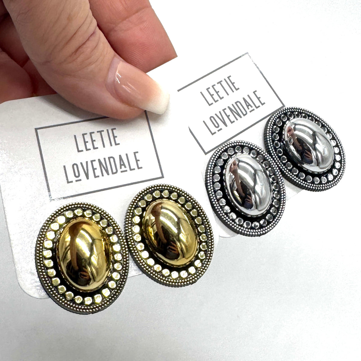 A close-up of a hand holding a white card printed with the brand name 'LEETIE LOVENDALE' above a pair of oval stud earrings. The card also shows a second pair of matching earrings.