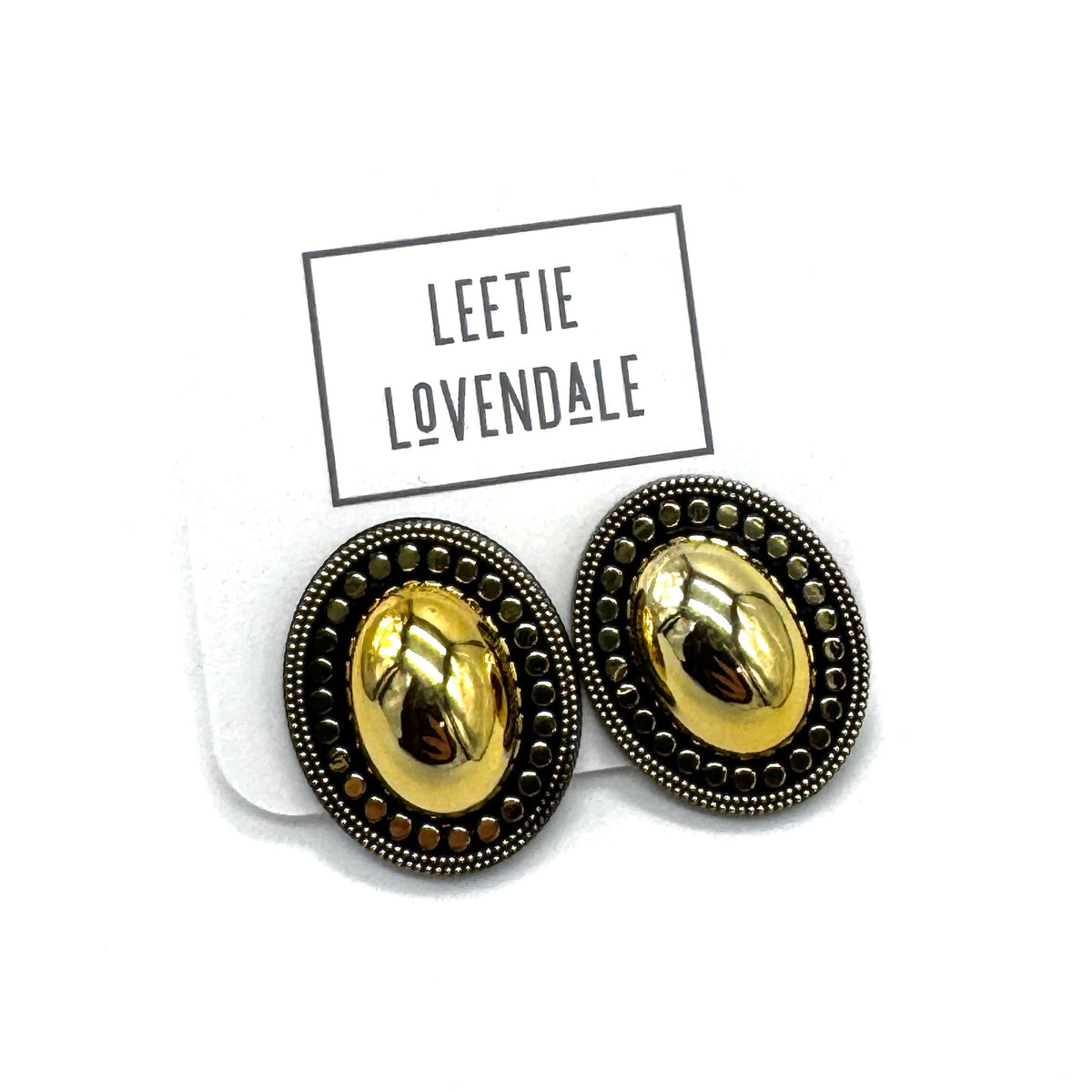 A pair of oval stud earrings with a shiny gold center and a dark bronze textured border, displayed on a white backing card with the brand name 'LEETIE LOVENDALE' printed above them.
