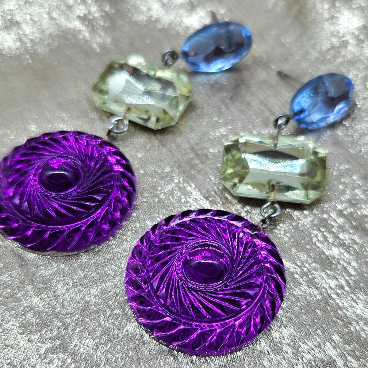 Purple circular earrings with blue and green shaped stones on a textured surface