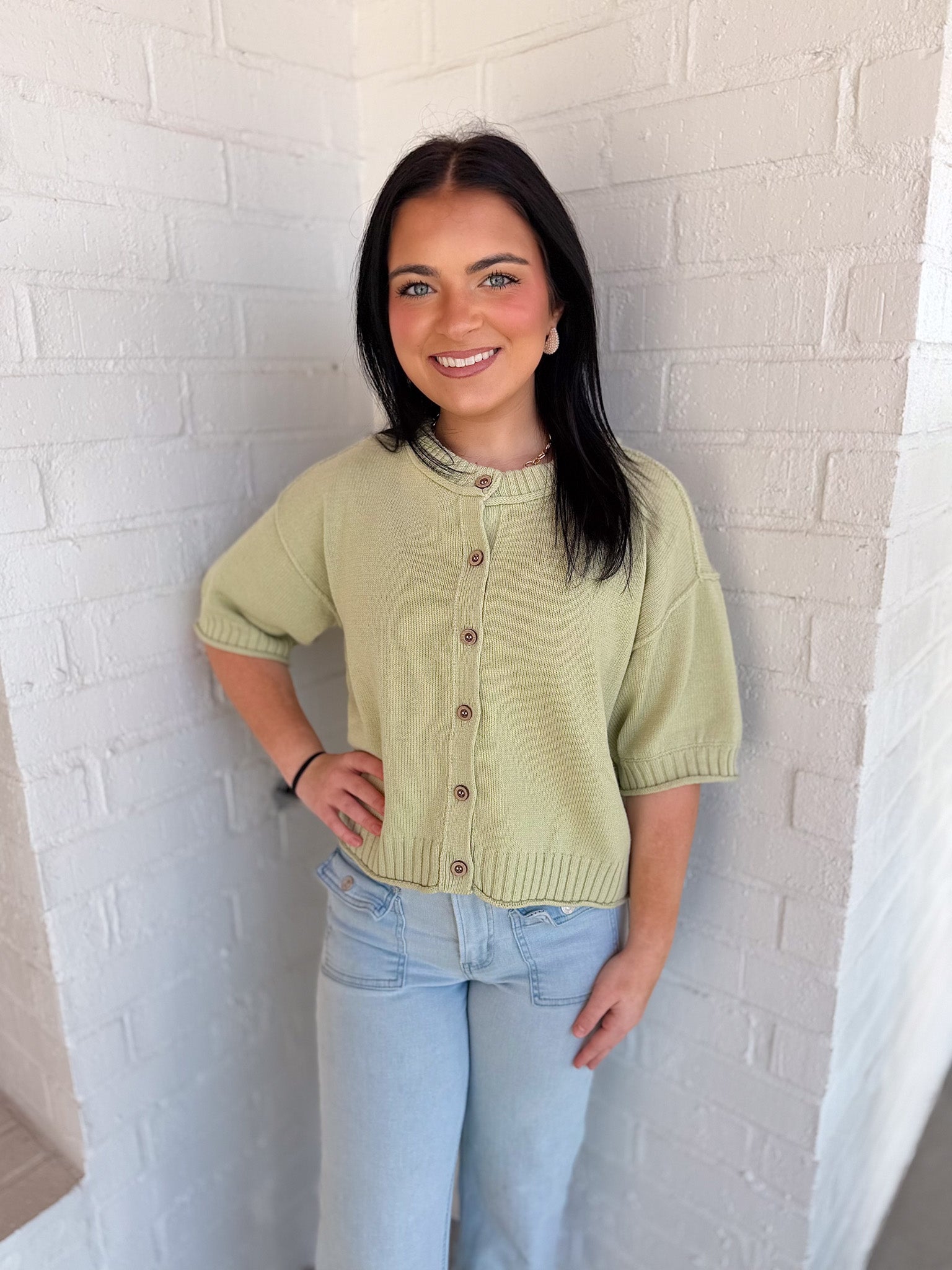 Woman wearing a light green knitted cardigan and light blue jeans against a white brick wall.