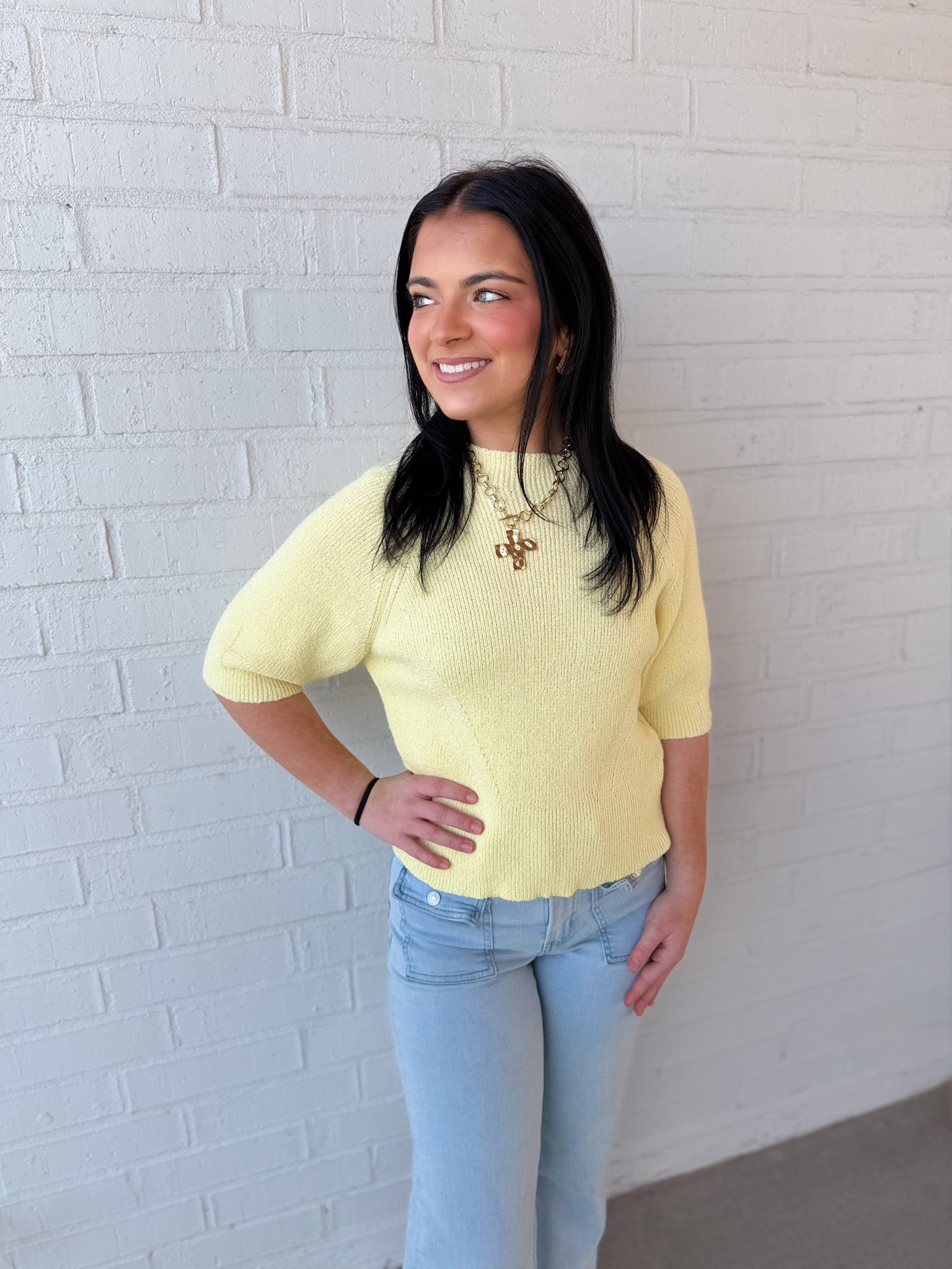 Woman wearing a yellow sweater and light blue jeans against a white brick wall.