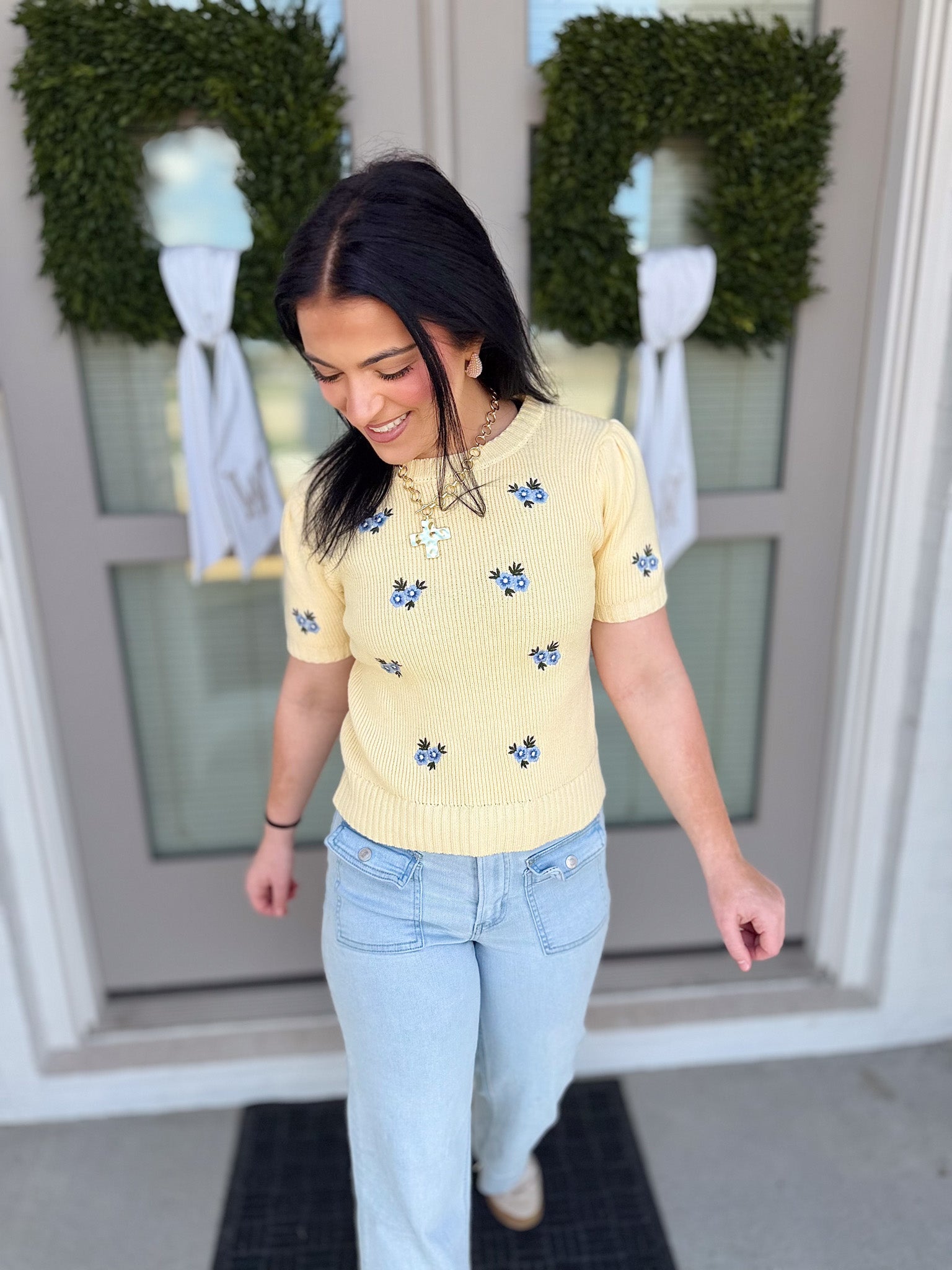 Woman wearing a yellow floral sweater and light blue jeans standing in front of a door with wreaths.
