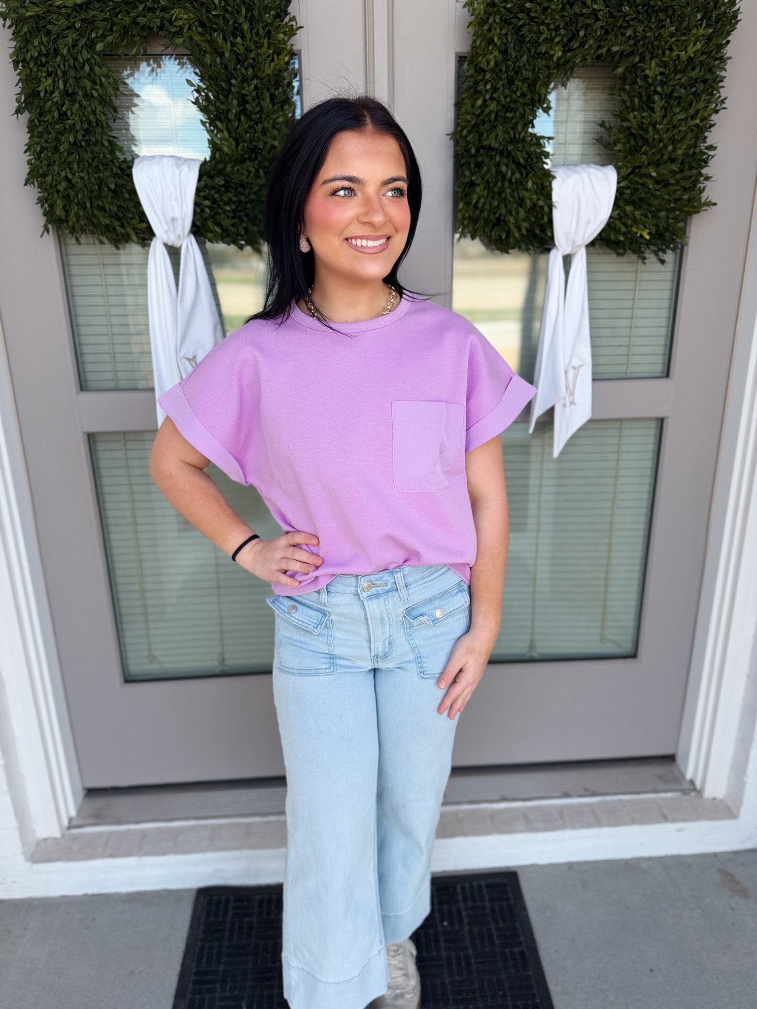 Woman wearing a purple shirt and light blue jeans standing in front of a door with wreaths.