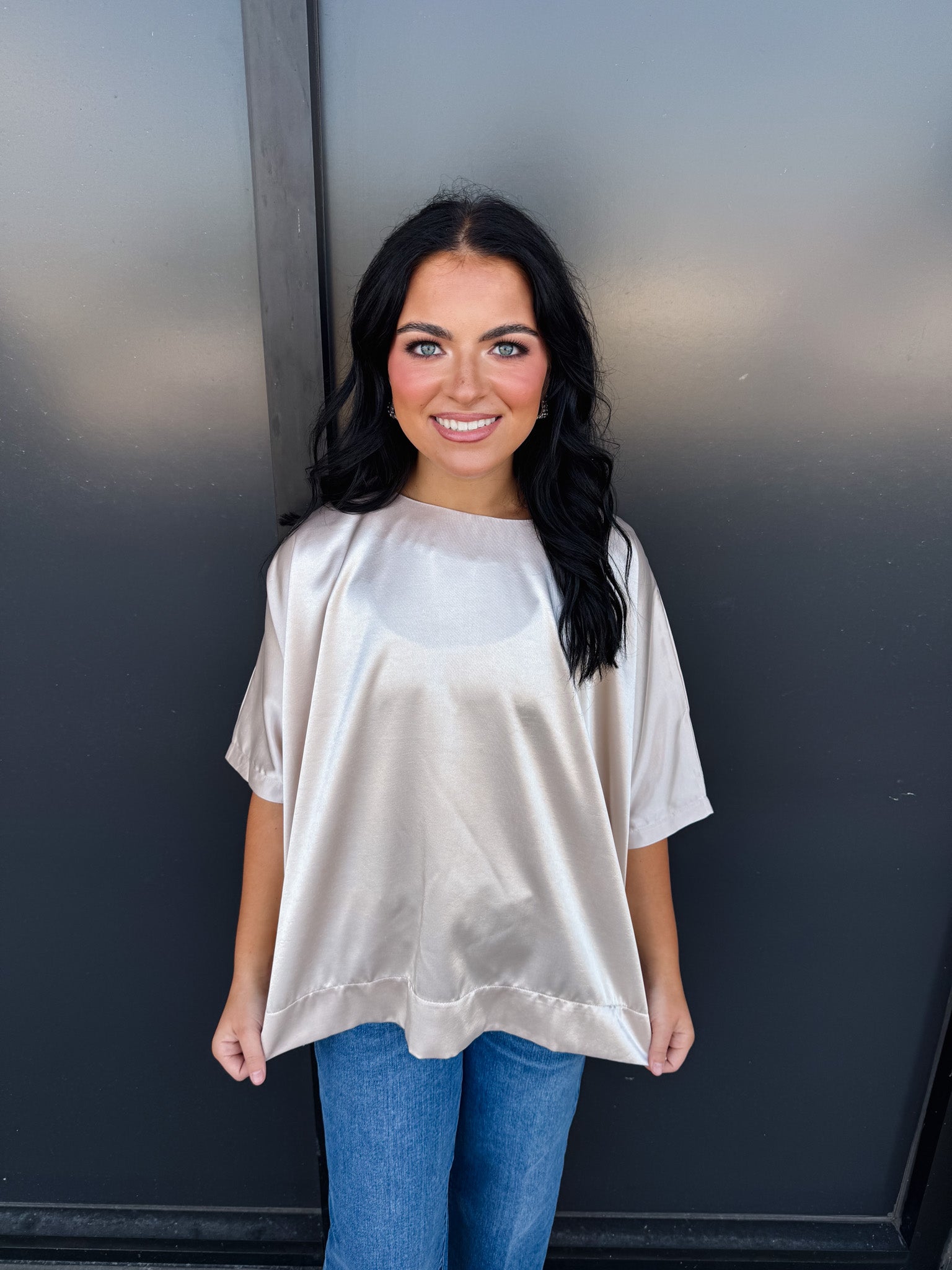 Woman wearing a beige oversized shirt and blue jeans against a dark wall.