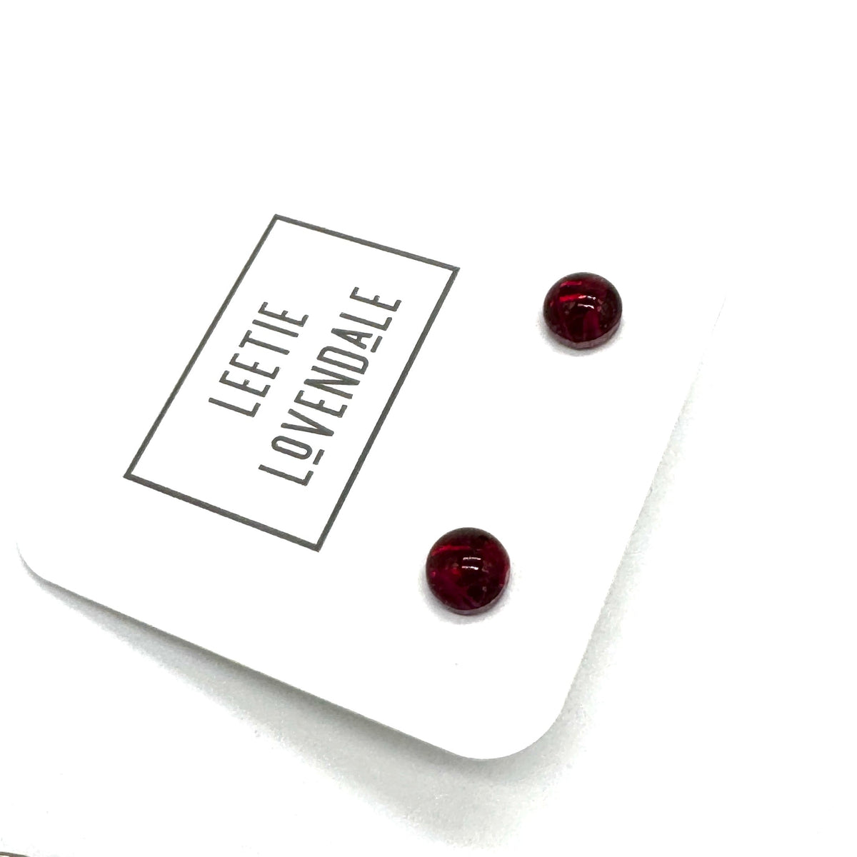 A pair of small round stud earrings in deep red resin, displayed on a white branded card that reads 'LEETIE LOVENDALE'.