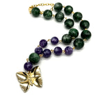A beaded necklace featuring dark green lucite beads, purple amethyst beads, and a textured gold butterfly pendant, displayed on a clean white background.